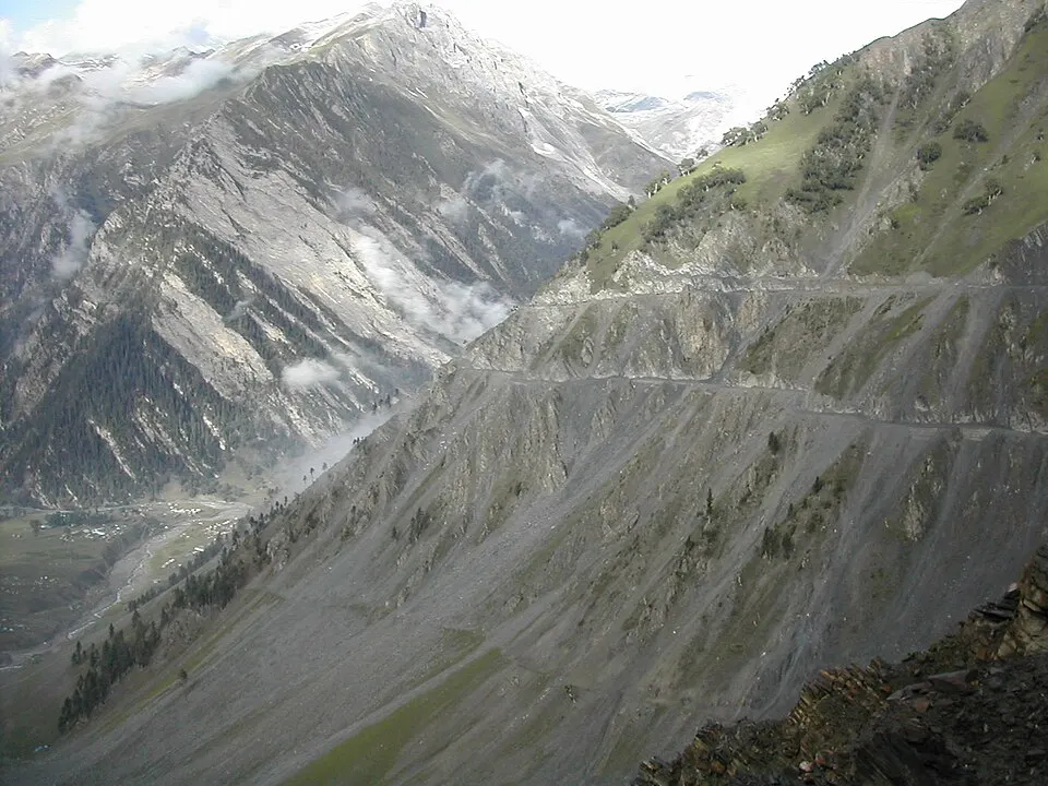 A narrow road cut along a steep, bare mountainside above a green valley with mist and snowy peaks.