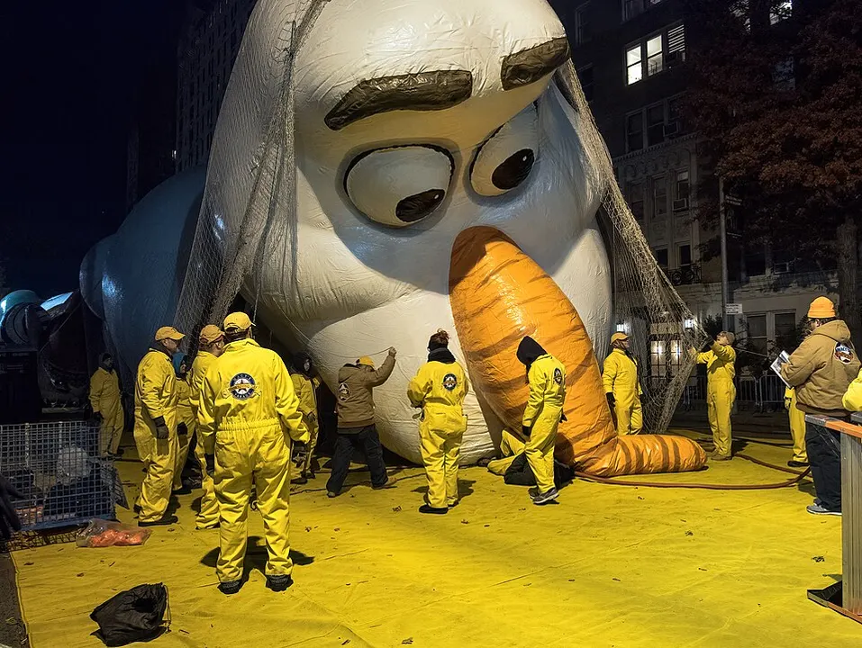 A huge inflatable balloon with a big orange nose and wide eyes being held down by workers in yellow jumpsuits on a city street at night.