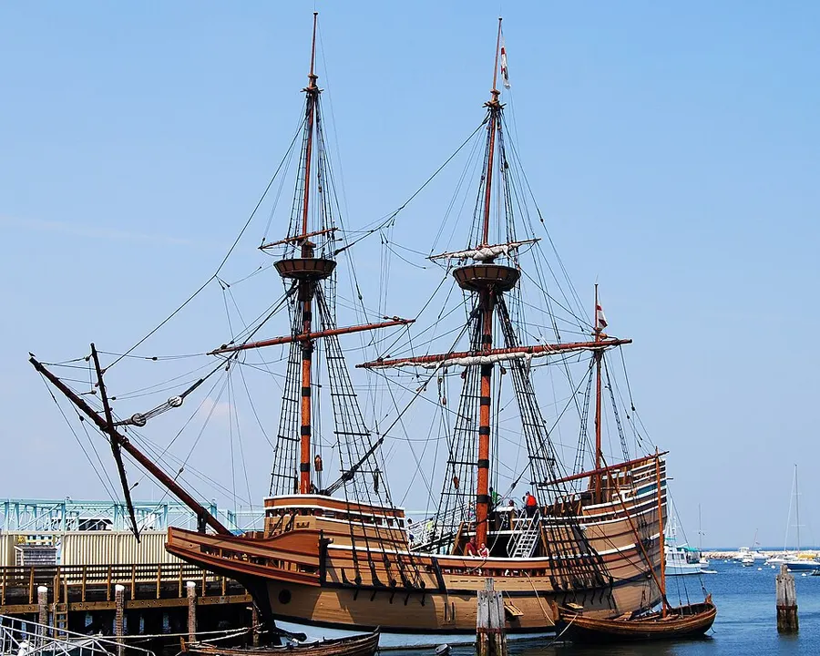 A wooden sailing ship with two tall masts and rigging moored at a dock on a sunny day.