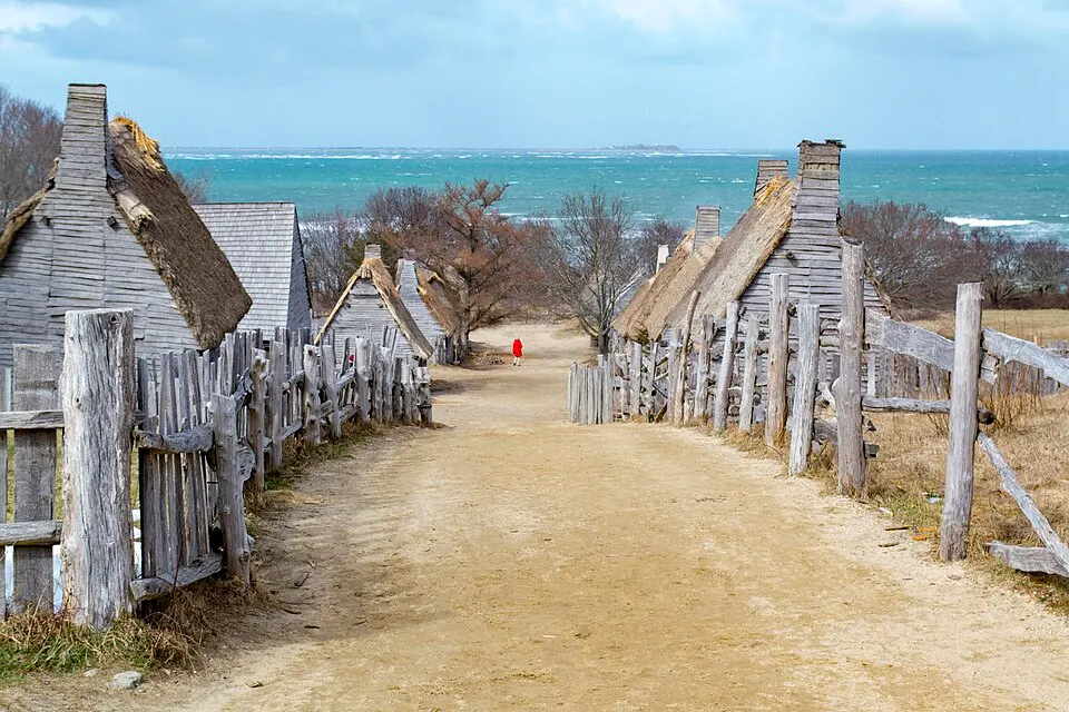 A dirt path between rough wooden fences and thatched, weathered wooden houses with the sea beyond.