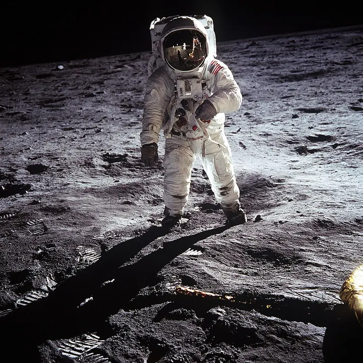 An astronaut in a white spacesuit and gold-visored helmet standing on grey, dusty ground with boot prints.