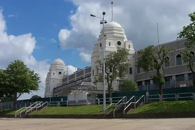 A large white stadium entrance with two domed towers rising above a walkway and grassy banks.