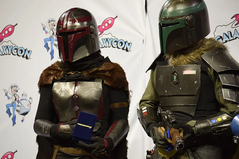 Two people in armoured space-warrior costumes and helmets, one in red and silver, one in green and black, stand in front of a convention backdrop.