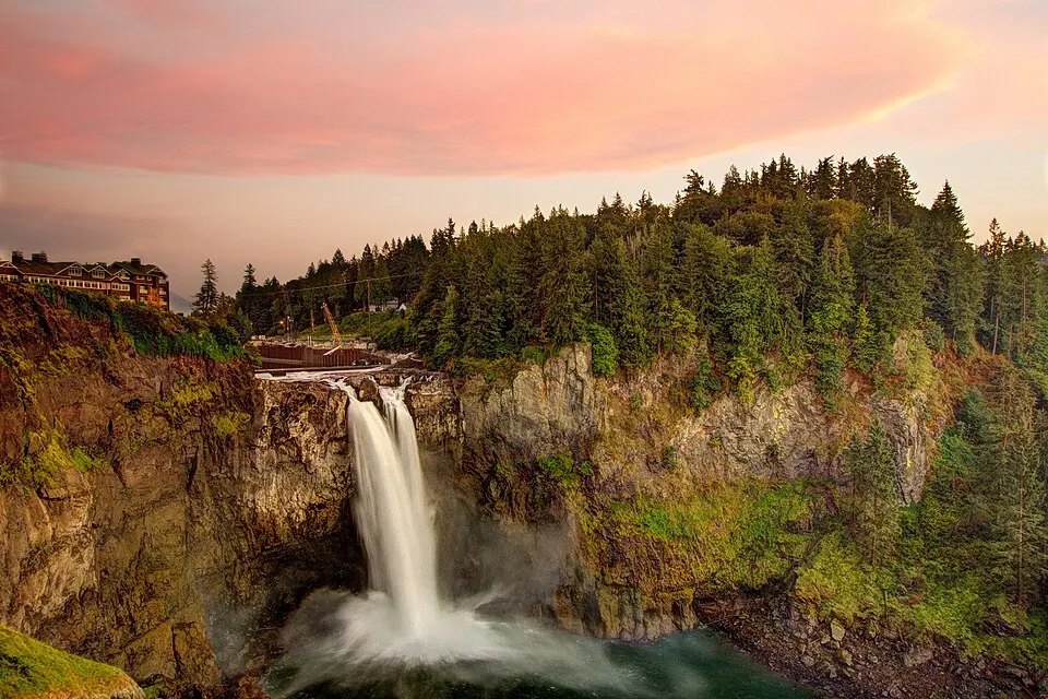 A tall waterfall drops into a misty pool below a forested cliff under a pink sunset sky.
