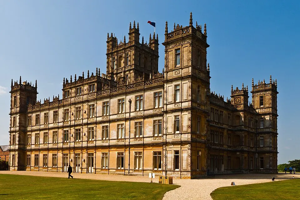 A grand sandstone country house with tall corner towers and many windows, beside a lawn under a blue sky.