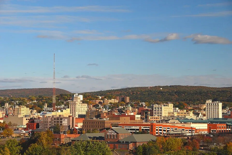 A town of brick buildings in front of tree-covered hills and a tall red radio mast under a blue sky.