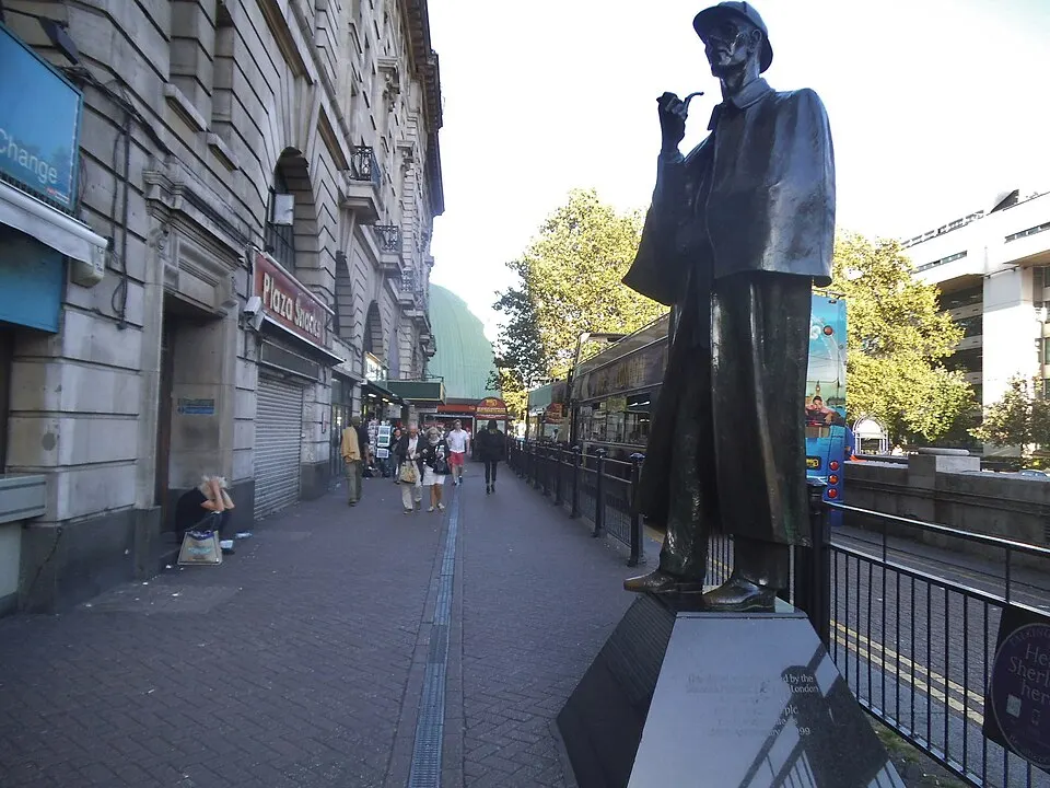 A bronze statue of a man in a deerstalker cap and cape holding a pipe, beside a busy London pavement.