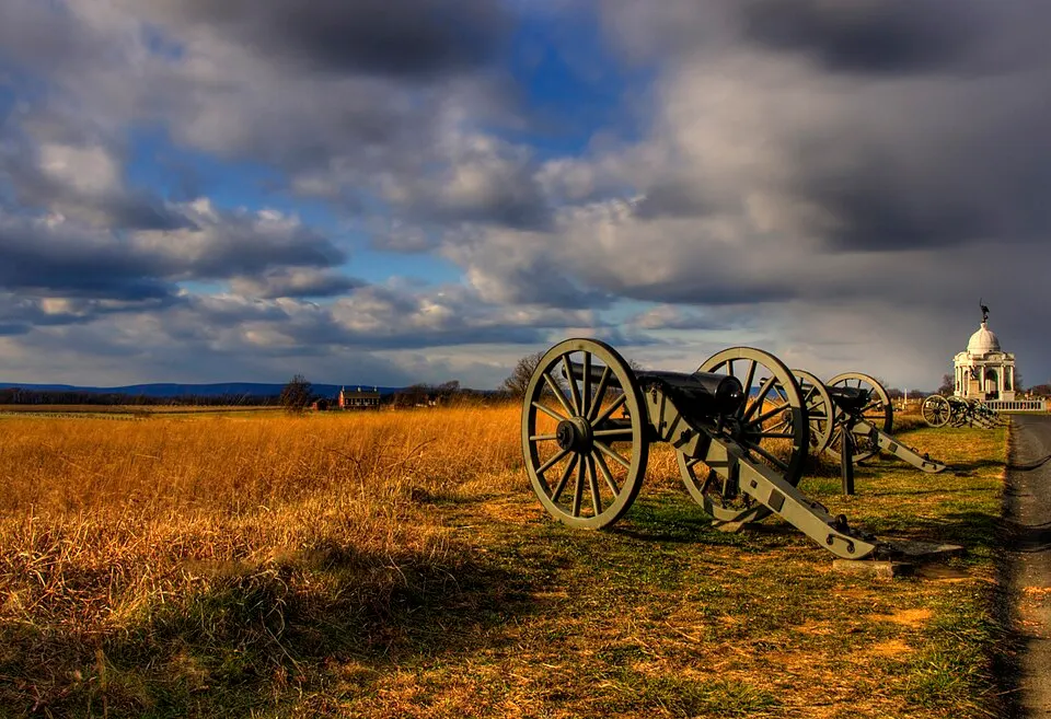 Old green cannons lined up along a grassy field under a dramatic cloudy sky, with a white monument in the distance.
