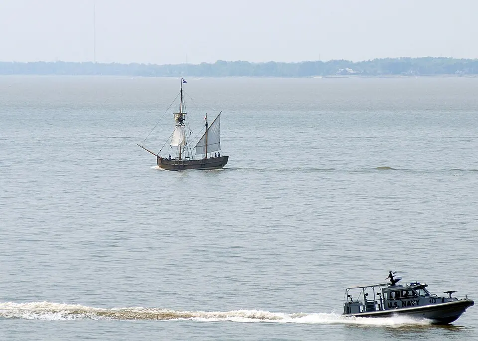 A small wooden sailing ship with square sails on a wide river, with a grey patrol boat speeding past in the foreground.