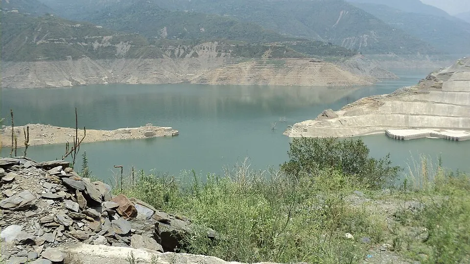 पहाड़ियों से घिरा एक विशाल जलाशय, किनारे पर कंक्रीट की संरचना और सामने चट्टानें तथा झाड़ियाँ।