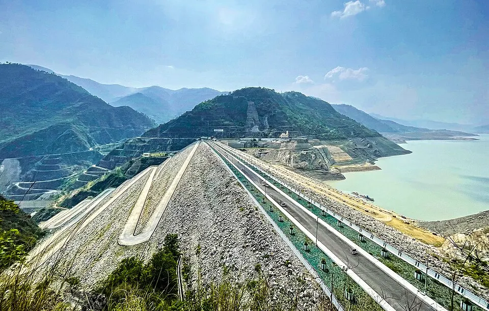 A huge rock-and-earth dam wall with a road along its crest holds back a pale green reservoir among steep hills.