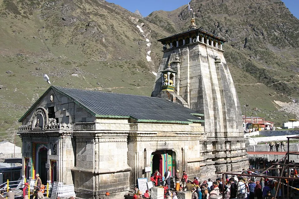 An old grey stone temple with a tall tower stands with pilgrims in front, on a bare mountain slope.