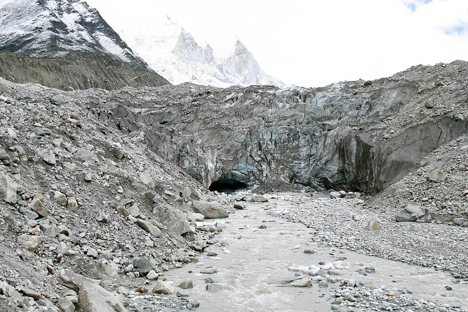 A grey ice cave opens at the bottom of a rubble-covered glacier wall, with a silty stream and snow peaks above.
