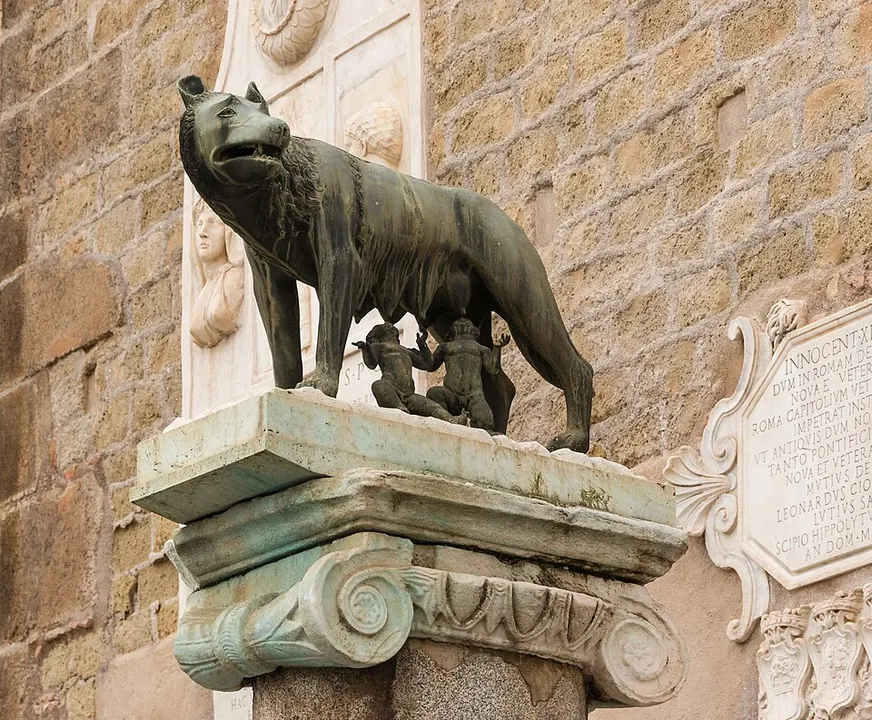 A bronze statue of a wolf with two small children beneath her, standing on a carved stone column against a stone wall.