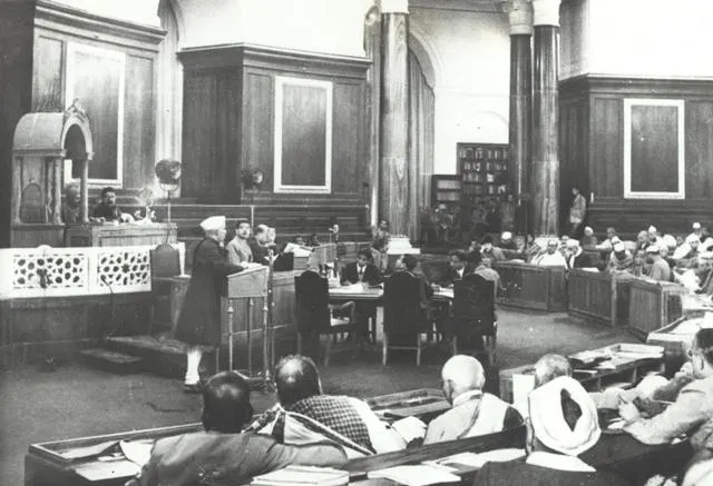 A black-and-white photo of a man in a cap speaking at a podium in a large wood-panelled assembly hall.