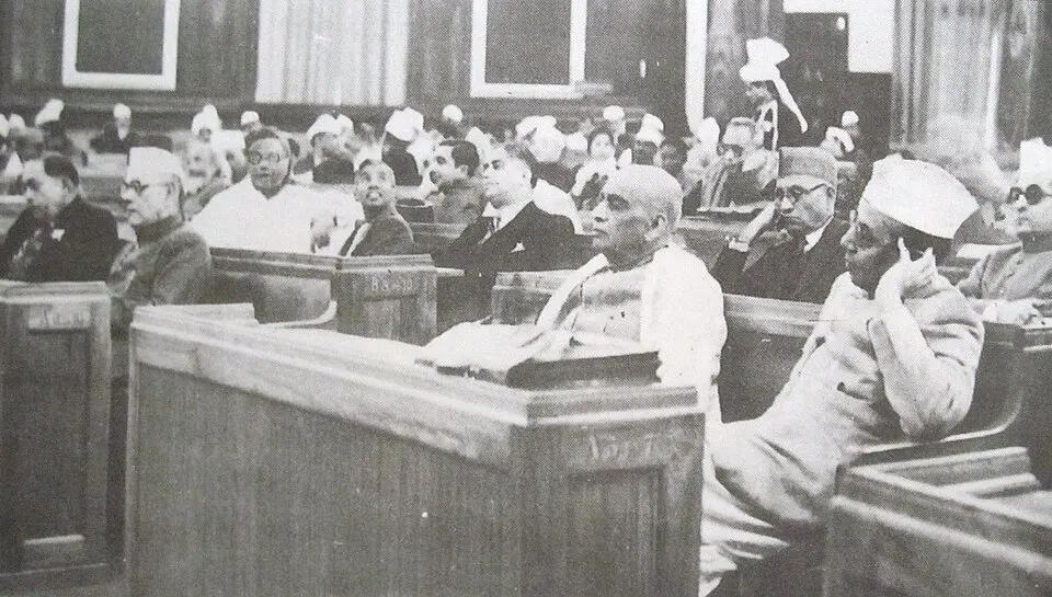A black-and-white photo of members in caps and white clothes seated at wooden desks in a crowded assembly hall.