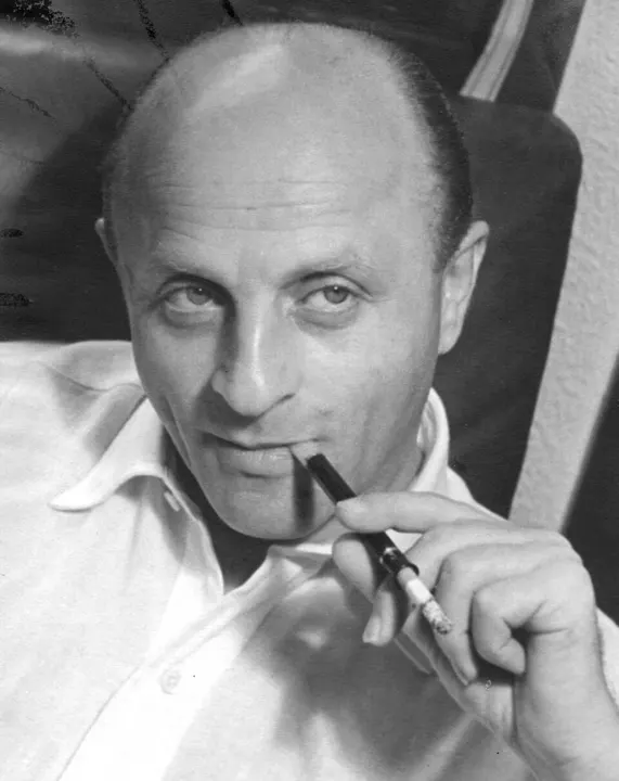 A black-and-white photo of a balding man holding a pen to his lips.