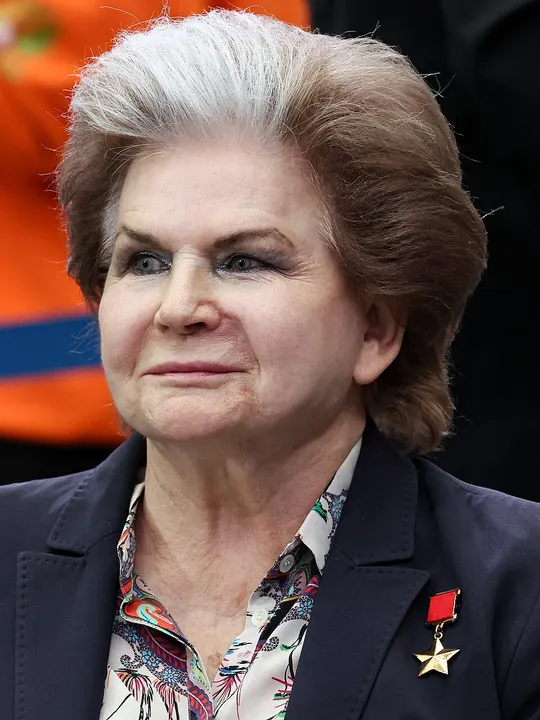 An elderly woman with silver hair in a dark jacket with a medal.
