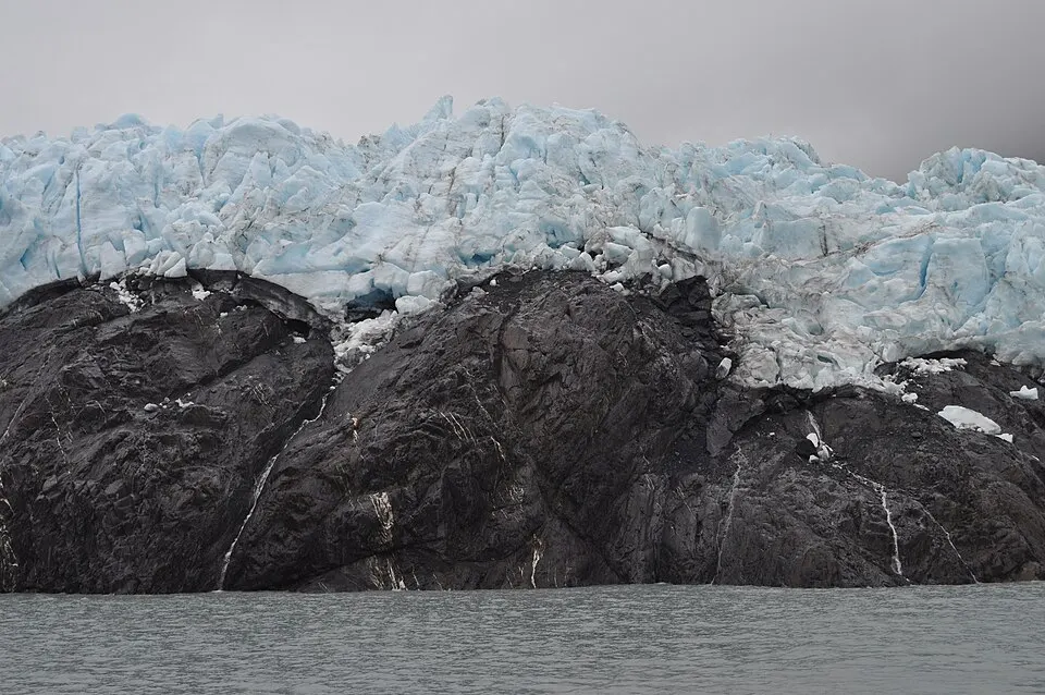 A wall of pale blue glacier ice above dark rock, rising from grey water.