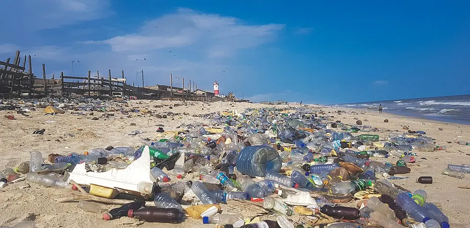A sandy beach covered with thousands of plastic bottles and rubbish, with the sea and a red-and-white lighthouse behind.
