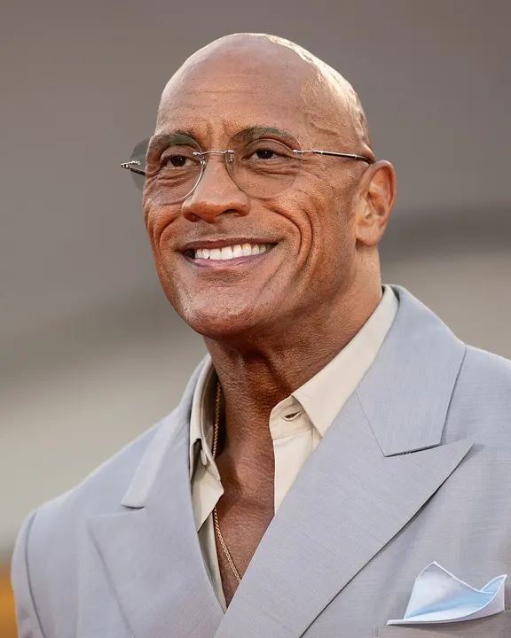 A muscular smiling bald man in a dark suit.